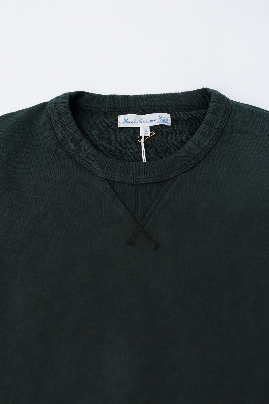 Close-up of Merz b. Schwanen forest green fleece sweatshirt front collar, ribbed neckline, V-stitch detail, and brand tag at Dant Indianapolis.