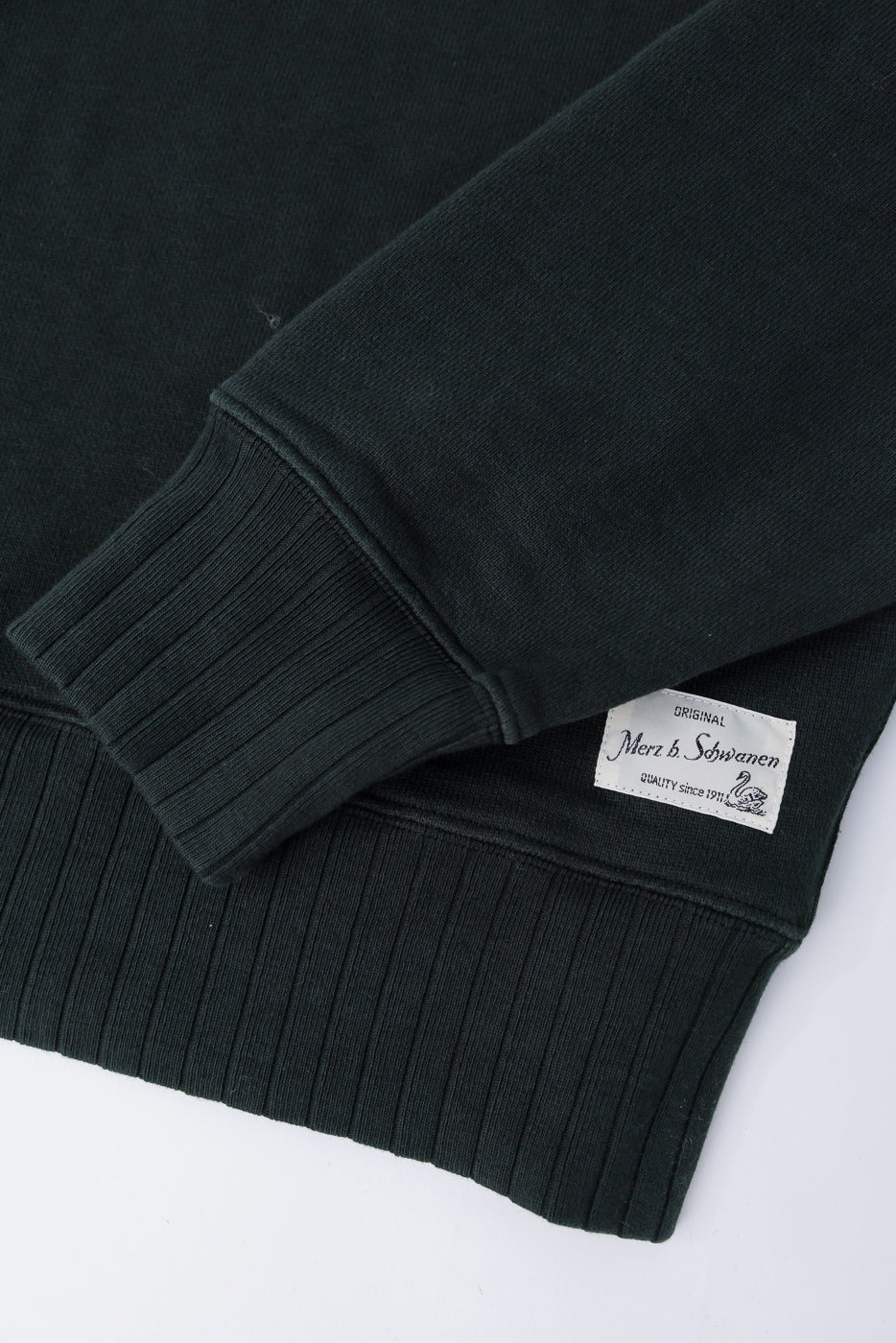 Close-up of Merz b. Schwanen forest green heavyweight fleece sweatshirt front hem, ribbed cuff, and logo tag, highlighting textured cotton fabric. No model shown.