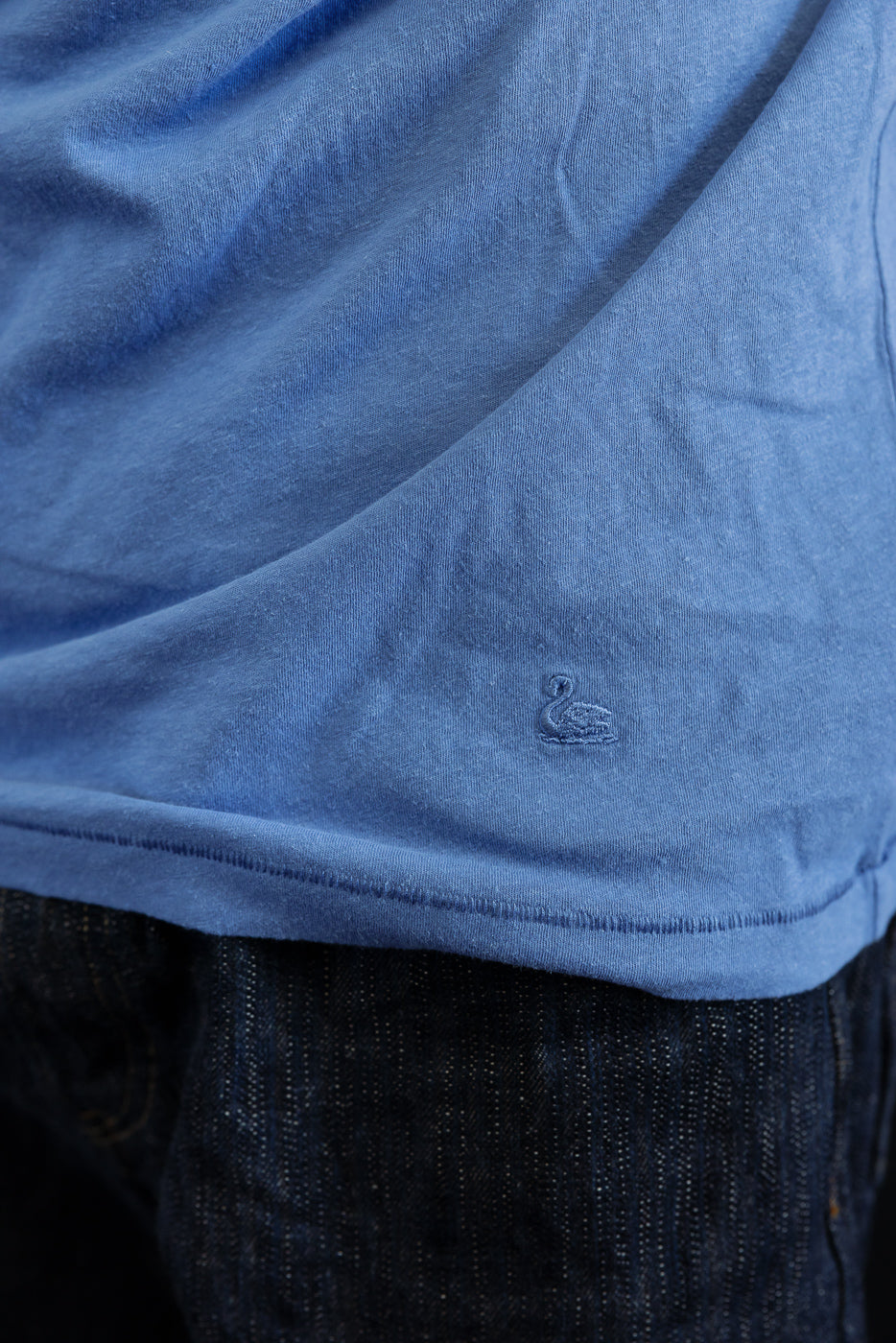 Close-up of faded blue cotton hemp t-shirt hem, left side, small swan embroidery detail.