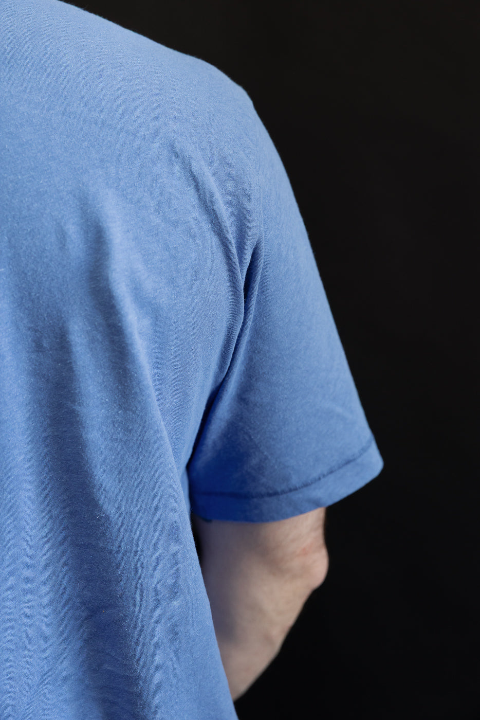 Close-up of male model’s upper right back and sleeve of blue cotton hemp T-shirt, fabric texture visible.
