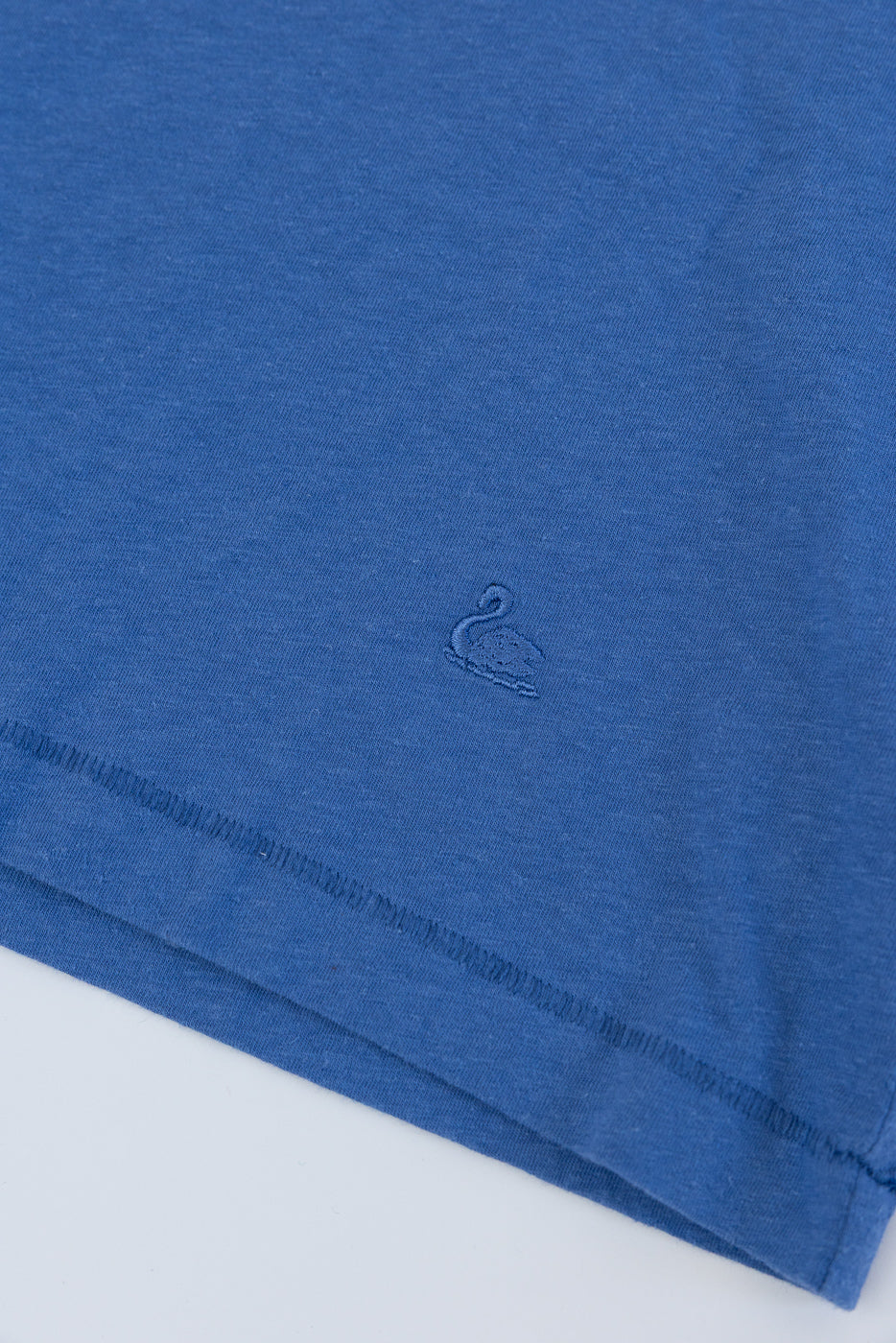 Close-up of front hem on faded blue cotton hemp t-shirt, showing embroidered swan logo and fabric texture.