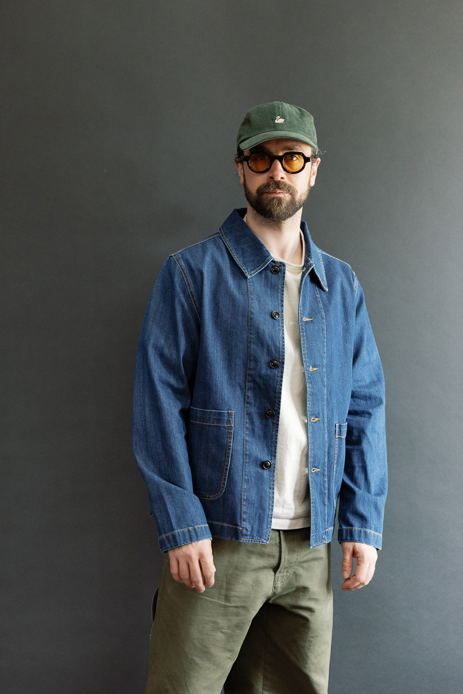 Man wearing JKT08DN.520MB - Cropped Worker's Denim Jacket, showcasing its classic collar and corozo nut button closure, styled over a white T-shirt, paired with workwear-style trousers and a baseball cap.