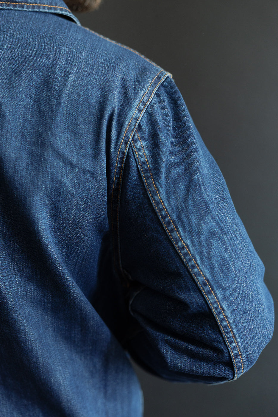 The JKT08DN.520MB Cropped Worker's Denim Jacket features rich blue organic cotton denim with orange stitching, showcasing a classic collar and detailed craftsmanship on the back and upper arm.