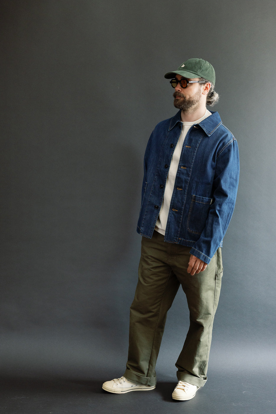 Man wearing Merz b. Schwanen JKT08DN Cropped Worker's Denim Jacket, showcasing a vintage dark wash with a cropped cut and classic collar, styled casually with workwear influence against a dark-gray backdrop.