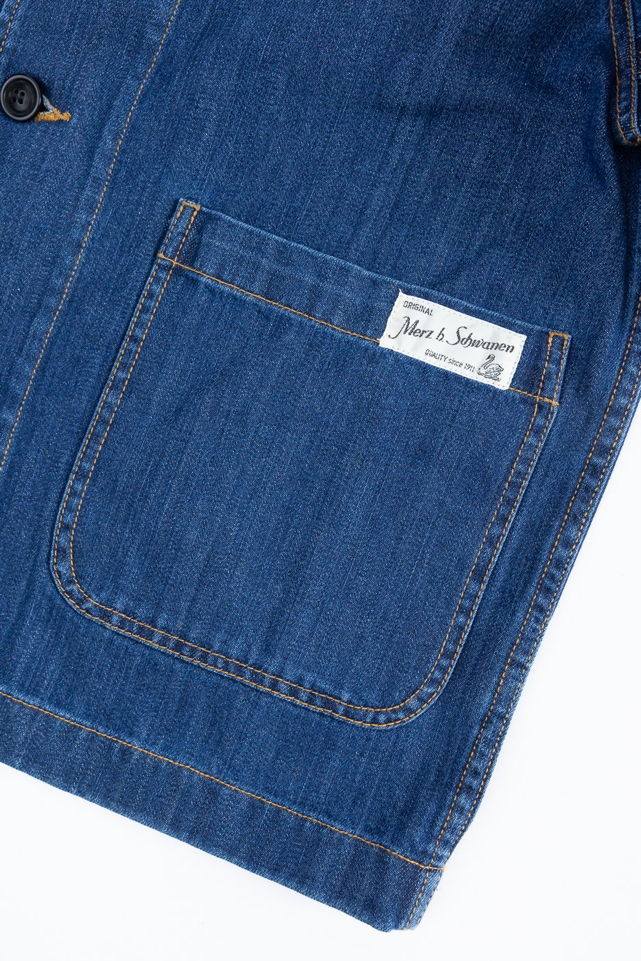 Close-up of the JKT08DN.520MB Cropped Worker's Denim Jacket, showcasing a blue denim patch pocket with orange stitching and a Original Merz b. Schwanen label, emphasizing detailed craftsmanship.