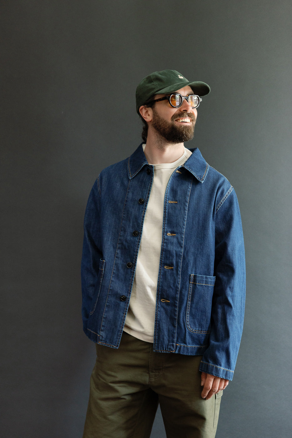 A bearded man models the JKT08DN.520MB Cropped Worker's Denim Jacket, showcasing its vintage dark wash, cropped cut, and classic collar, styled with tortoiseshell glasses and a baseball cap.