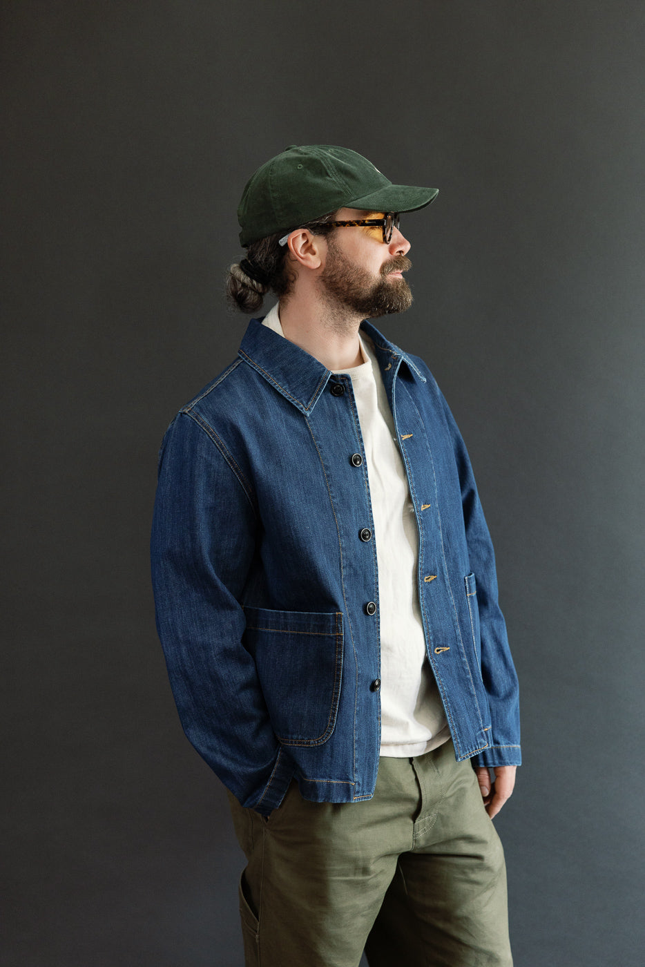 Man wearing the JKT08DN.520MB Cropped Worker's Denim Jacket, featuring a vintage dark wash, classic collar, corozo nut buttons, and triangular underarm inserts, styled with olive-green pants and a baseball cap.