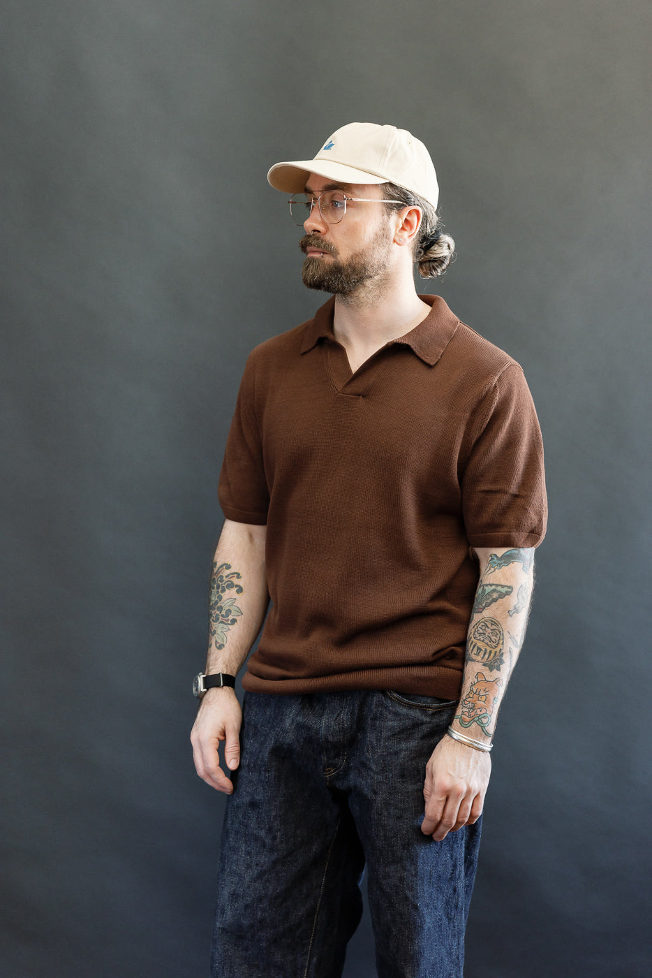 A man models the Merz b. Schwanen KNITPOLO02.192.7 Chocolate Polo, featuring a soft, relaxed knit and small open V-neck, paired with dark-wash denim, showcasing detailed tattoos.