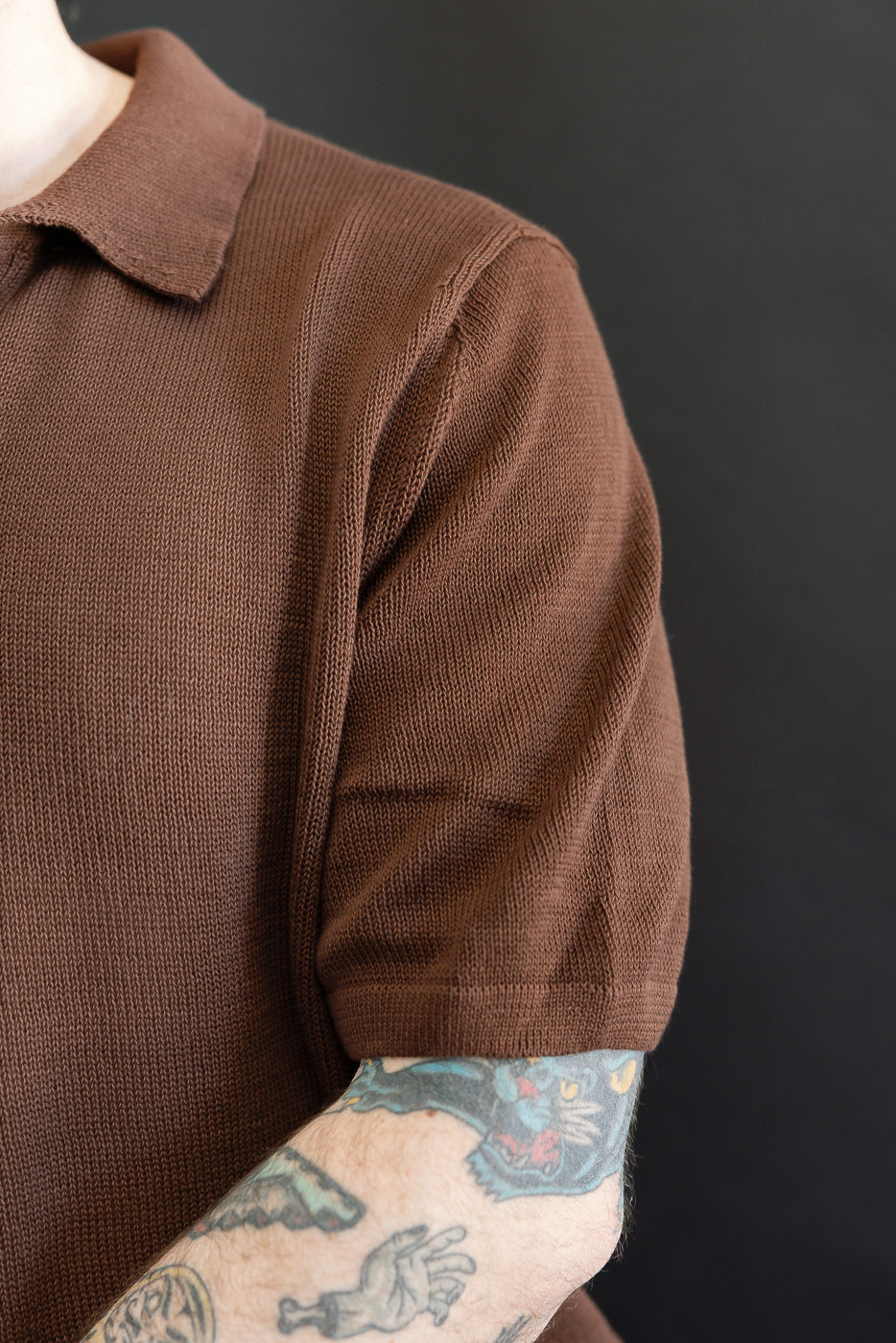 Close-up of someone wearing the KNITPOLO02.192.7 - Cotton Short Sleeve Polo Shirt, All Plant Based - Chocolate, showcasing its soft knit texture and relaxed V-neck, with colorful tattoos visible on the forearm.