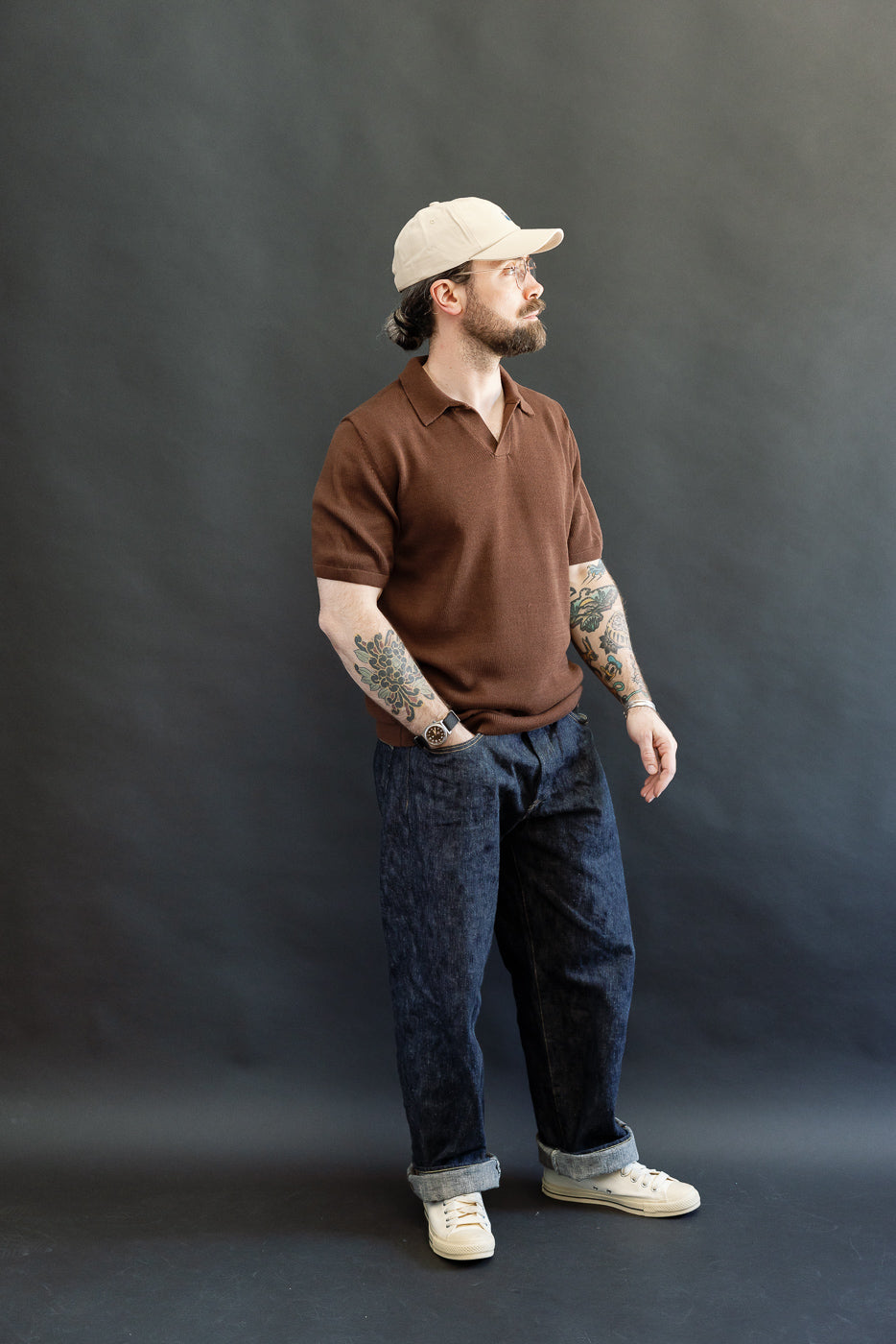 Man modeling the KNITPOLO02.192.7 Cotton Short Sleeve Polo Shirt, Chocolate, with a soft knit texture and small V-neck, paired with cuffed denim, showcasing a relaxed, heritage-inspired style.