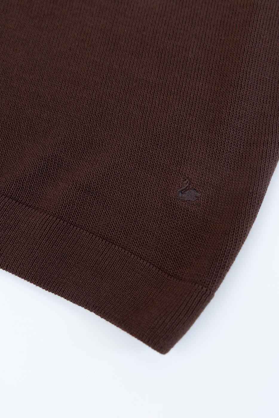 Close-up of the KNITPOLO02.192.7 - Cotton Short Sleeve Polo Shirt, All Plant Based - Chocolate, highlighting the ribbed hem with a subtle swan embroidery in matching thread, showcasing its knit texture.