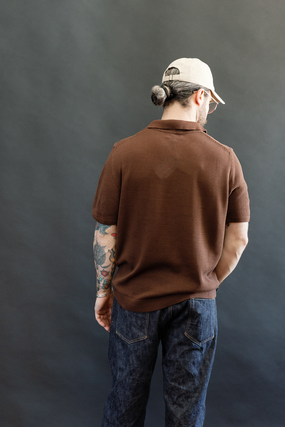 Person viewed from behind wearing the KNITPOLO02.192.7 - Cotton Short Sleeve Polo Shirt, All Plant Based - Chocolate, showcasing its relaxed knit texture and small V-neck, paired with dark blue denim jeans.