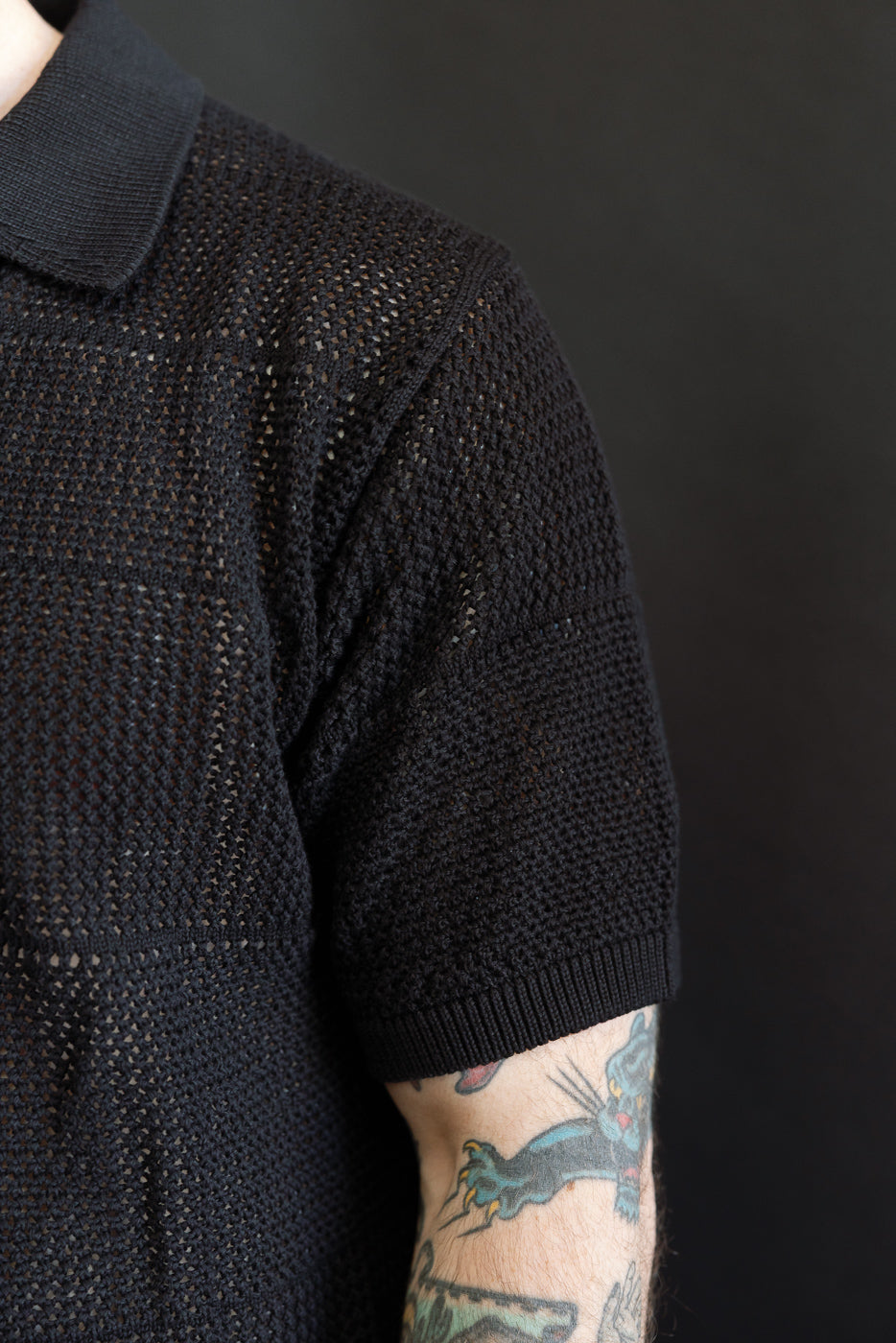 Close-up of male model’s right shoulder wearing deep black crochet cotton short sleeve knit shirt, showing textured fabric, ribbed sleeve hem, and collar.