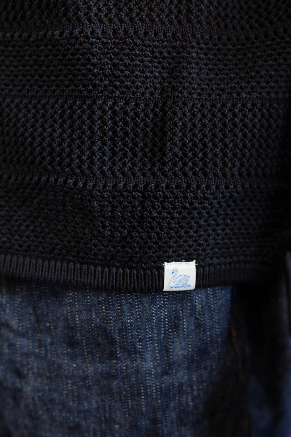 Close-up of men's deep black crochet cotton knit shirt hem with tag, front detail view.