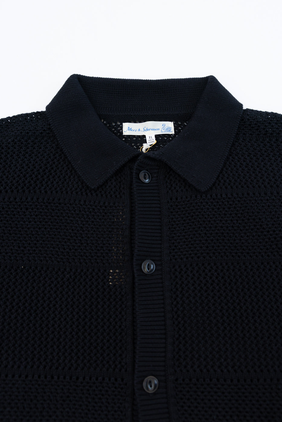 Close-up of the front upper section of a deep black crochet cotton short sleeve shirt, showing knit texture, ribbed collar, front buttons, and fabric tag.