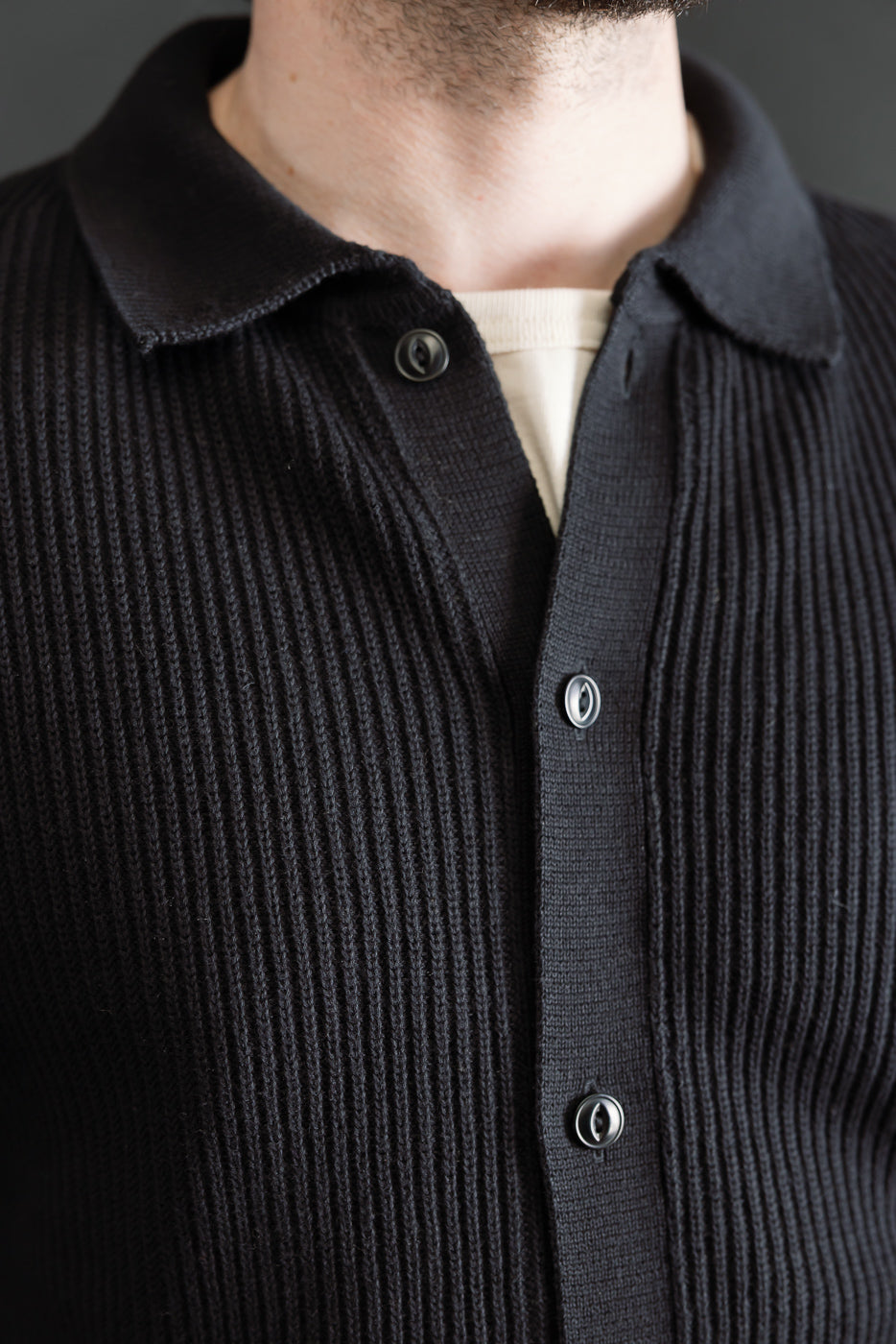 Close-up front of ribbed black cotton knit shirt on male model 5'9, 165lbs, athletic build, showing textured fabric, buttons, and collar.