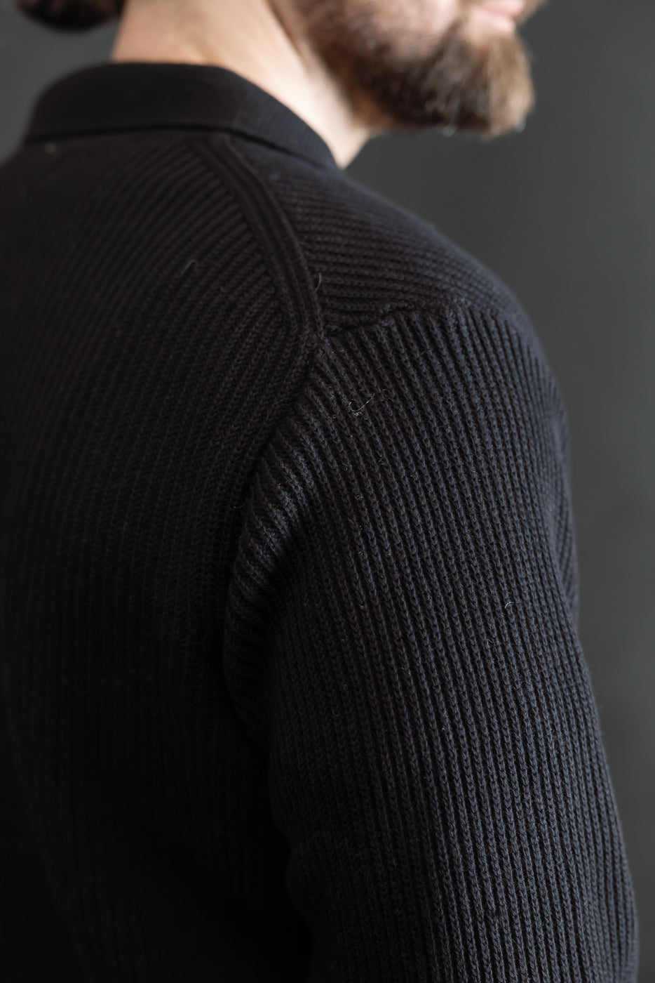 Close-up of male model's back right shoulder wearing ribbed black cotton long sleeve shirt, showcasing textured fabric and collar detail. Model is 5'9