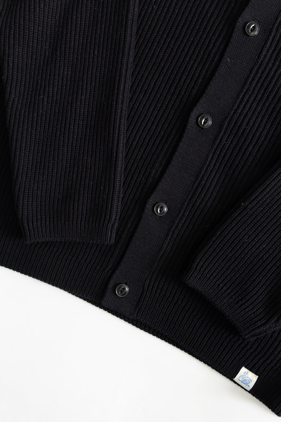 Close-up of deep black ribbed cotton long sleeve shirt, front hem, sleeve cuff, front buttons, visible fabric texture and woven tag detail.
