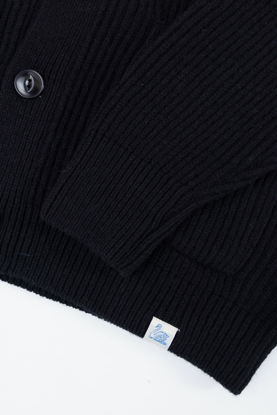 Close-up of the front bottom hem and sleeve of a black ribbed cardigan with a button and tag.