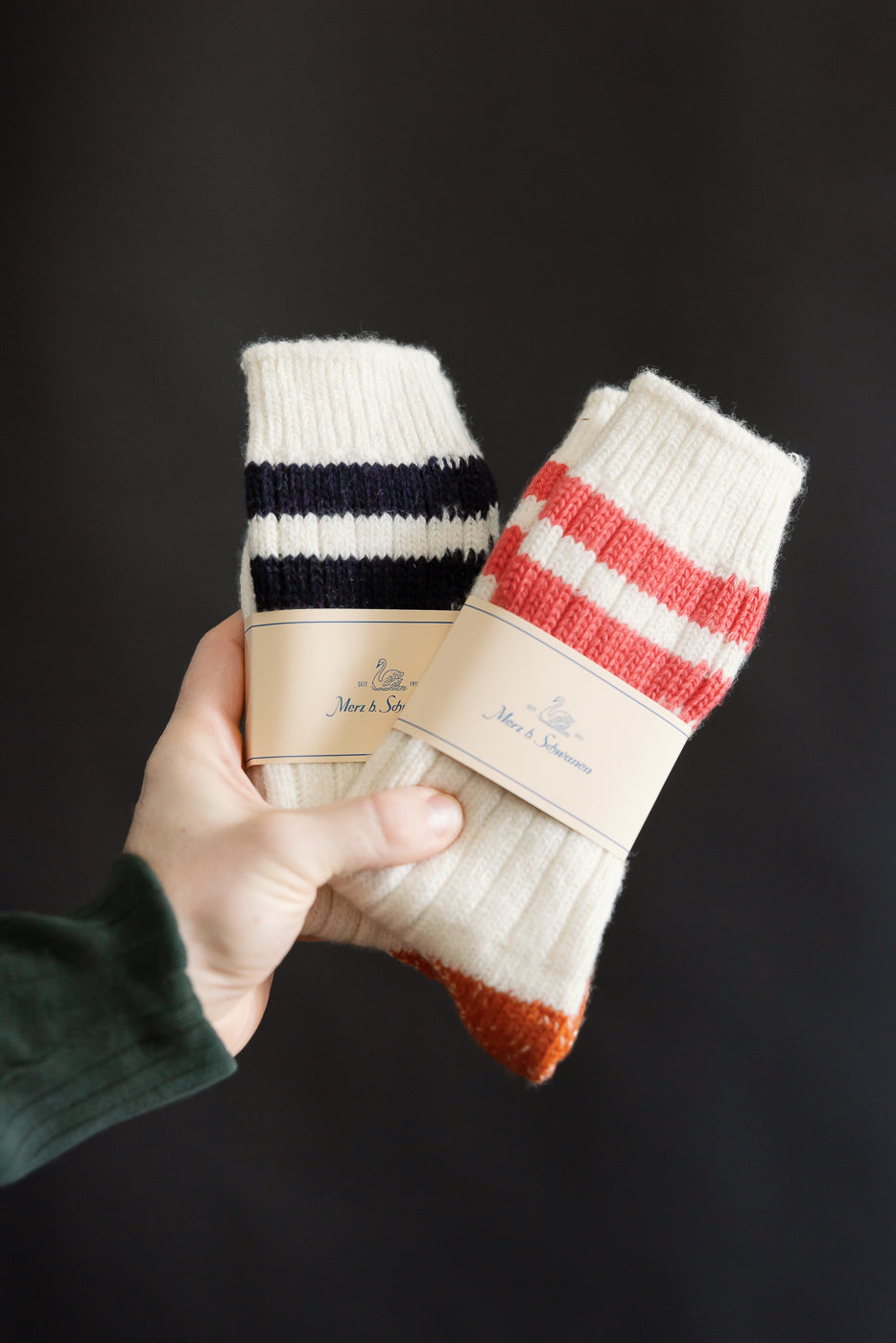 Close-up of two pairs of Merz b. Schwanen extra fine merino wool striped socks in Nature/Ink and Nature/Red colorways, showcasing ribbed knit cuffs and front packaging, photographed against a dark background for Dant contemporary casual apparel.