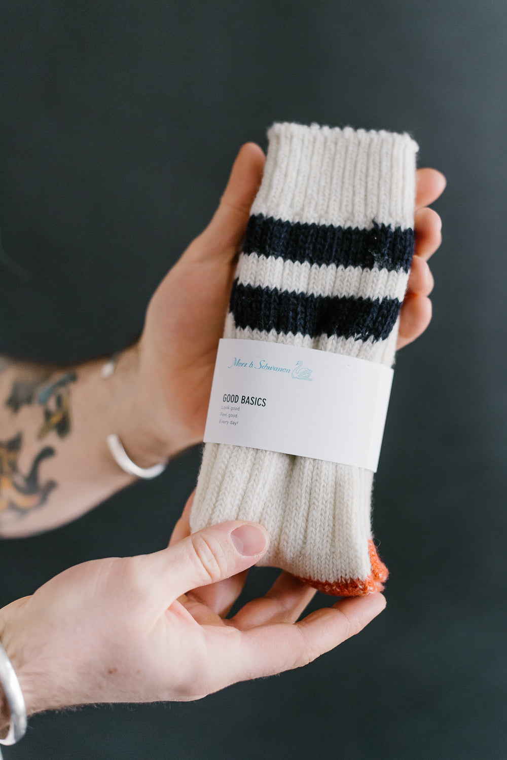 Close-up of Merz B. Schwanen extra fine merino wool socks, nature ink color, front view with visible label, held by tattooed male hand. Ribbed knit with navy stripes and orange toe detail.