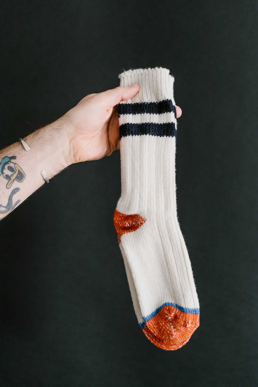 Close-up front view of Merz B. Schwanen extra fine merino wool sock in white with black stripes, orange heel and toe, held by male model's hand. Dant contemporary casual apparel.