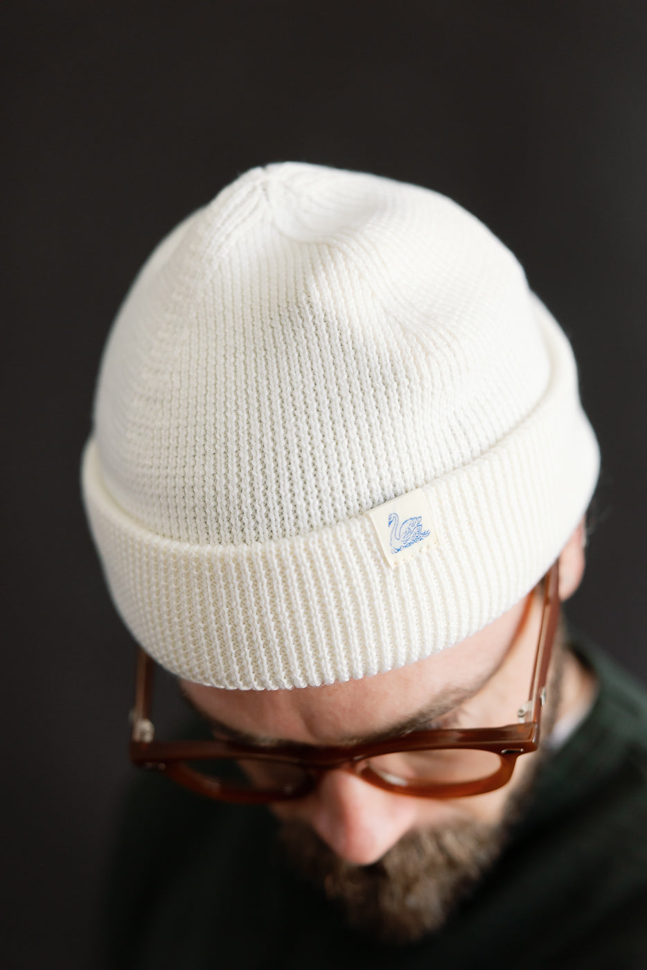 Close-up of male model wearing Merz B. Schwanen ribbed white merino wool watch cap, front view showing texture and logo tag, 5'9