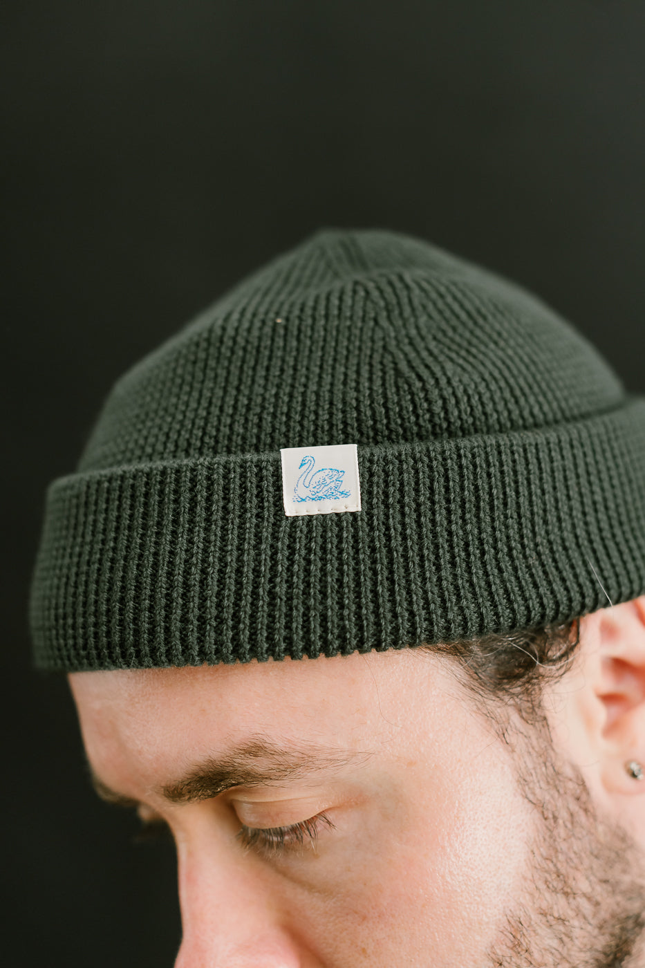 Close-up of ribbed Merz b. Schwanen army green merino wool watch cap, front logo tag, on male model 5'9