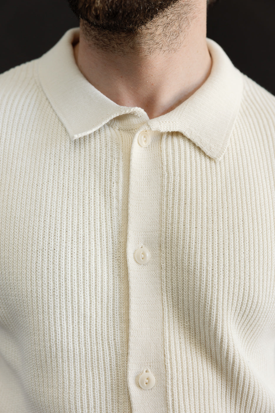 Close-up front view of ribbed cotton knit short sleeve shirt in natural color on male model, showing collar, buttons, and textured fabric. Model is 5'9