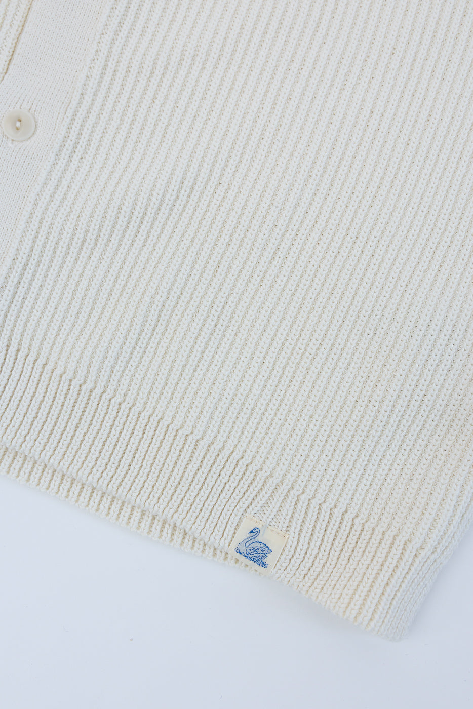 Close-up of front hem and side of natural ribbed cotton knit short sleeve shirt showing fabric texture, button, and swan tag detail.