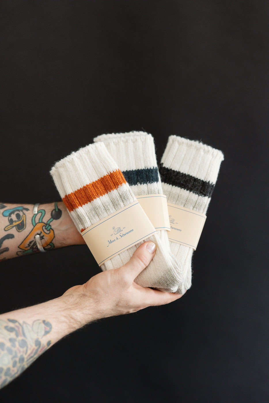 Close-up of three pairs Merz b. Schwanen recycled wool socks, ribbed knit, nature and amber color, front view in male model's tattooed hands.