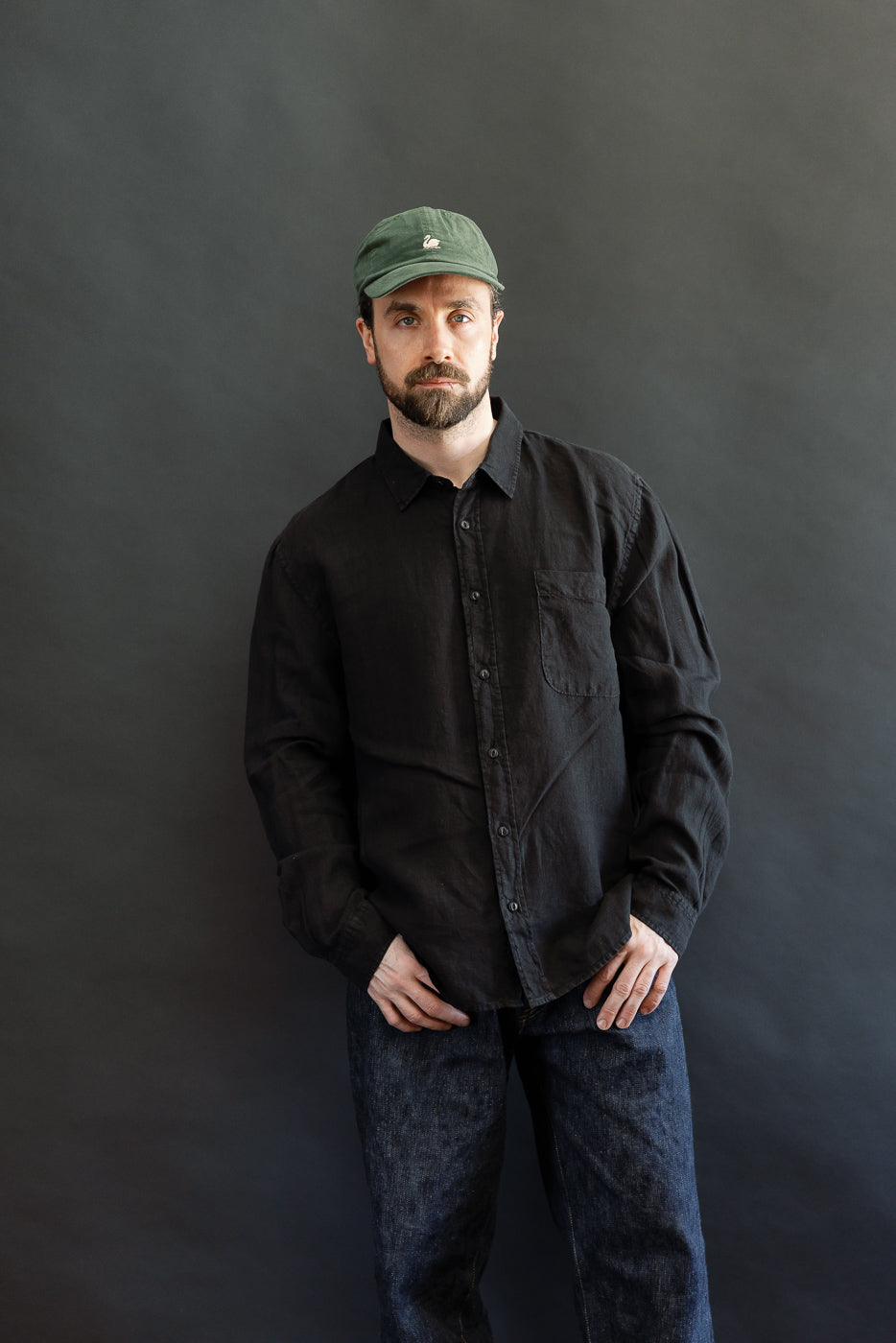 Man wearing the SHIRT20LI.99 - Organic Linen Long Sleeve Shirt - Black, featuring a chest pocket and relaxed fit, paired with dark blue jeans and a dark green baseball cap.