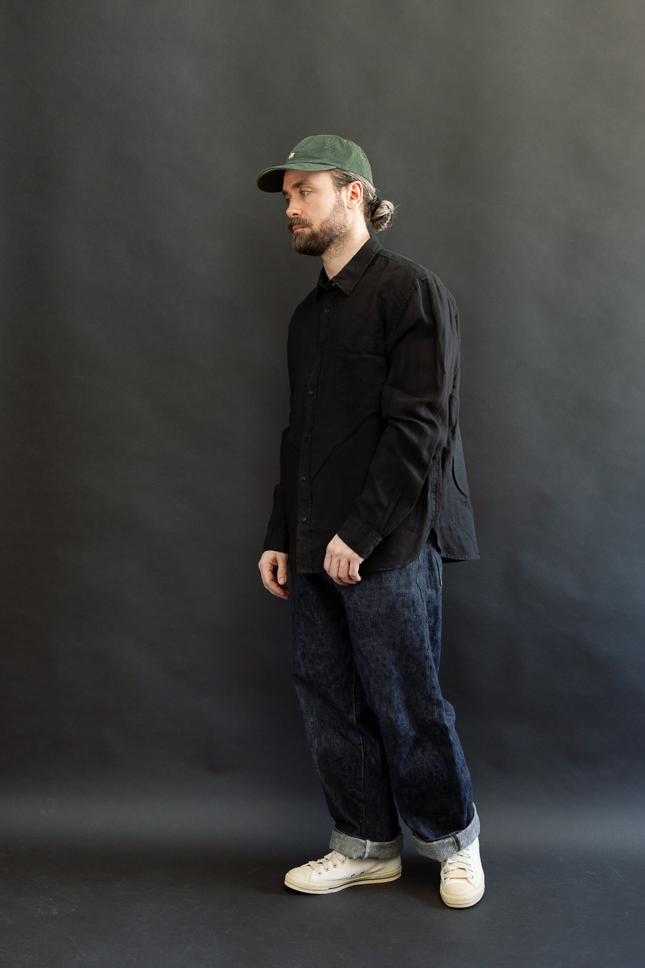 Bearded man in SHIRT20LI.99 - Organic Linen Long Sleeve Shirt, black button-up, relaxed fit, styled with rolled-up dark denim jeans and white sneakers, exemplifying Dant's heritage-inspired menswear.