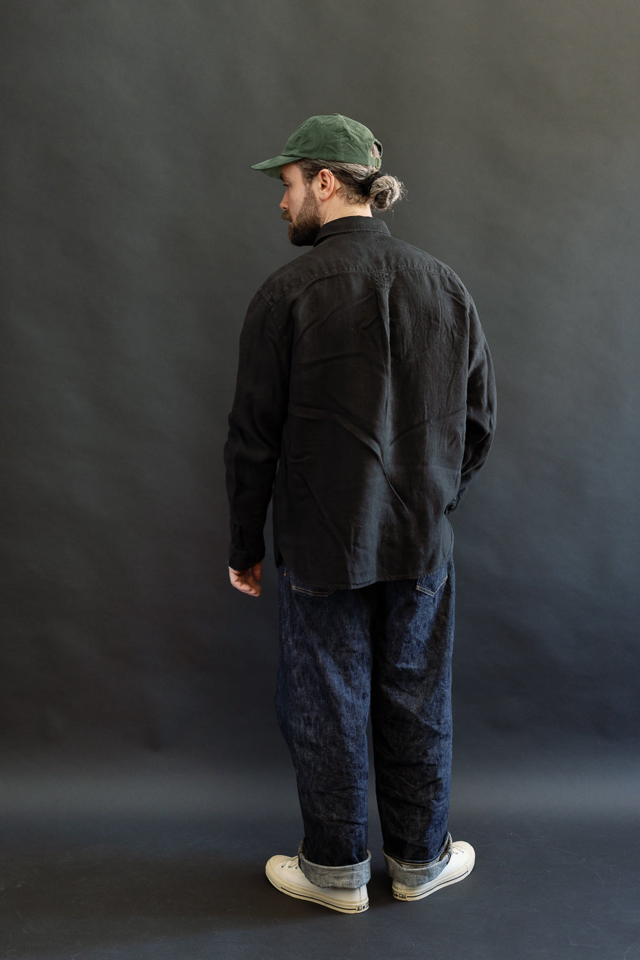 A person models the SHIRT20LI.99 - Organic Linen Long Sleeve Shirt - Black, showcasing its airy, cropped fit, button-up design, and box pleat, paired with cuffed jeans and white sneakers.