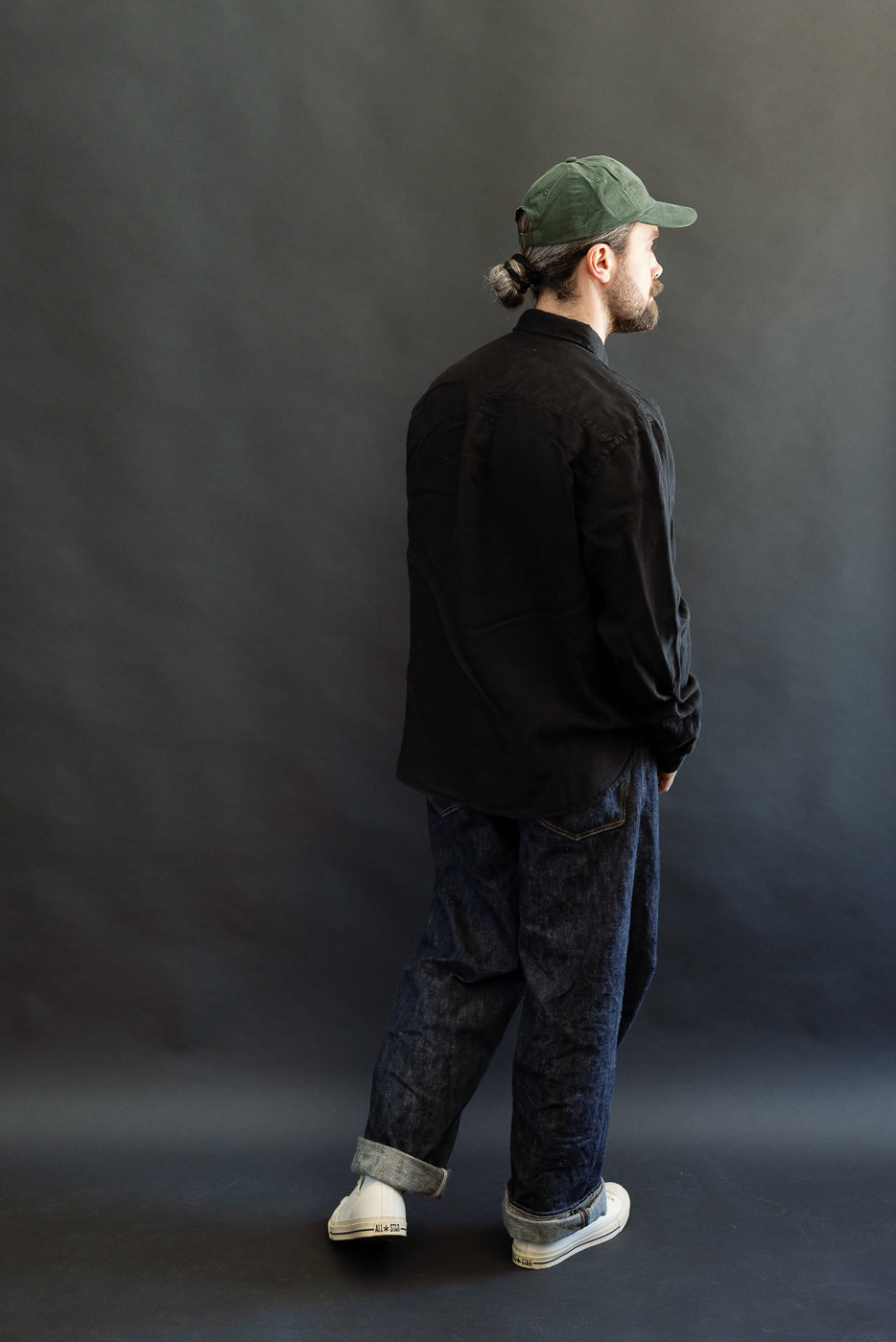 A man models the SHIRT20LI.99 - Organic Linen Long Sleeve Shirt - Black, showcasing its relaxed fit, rear pleat, and side slits, paired with denim jeans and white sneakers.
