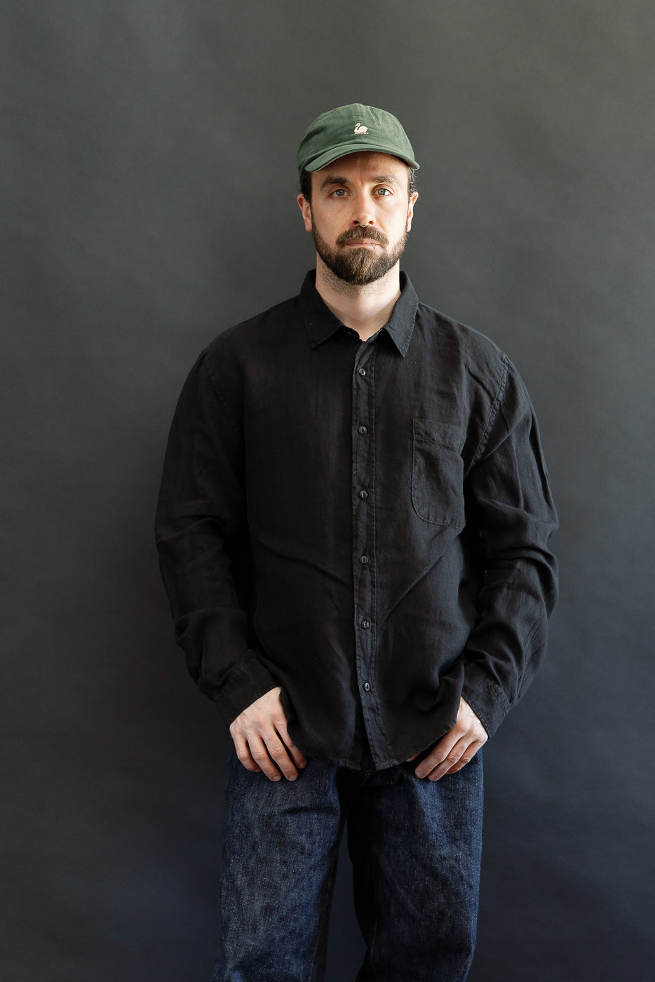 The SHIRT20LI.99 - Organic Linen Long Sleeve Shirt - Black, worn by a bearded man, showcases a relaxed fit with a swan-embroidered chest pocket, corozo nut buttons, and side slits for ease.