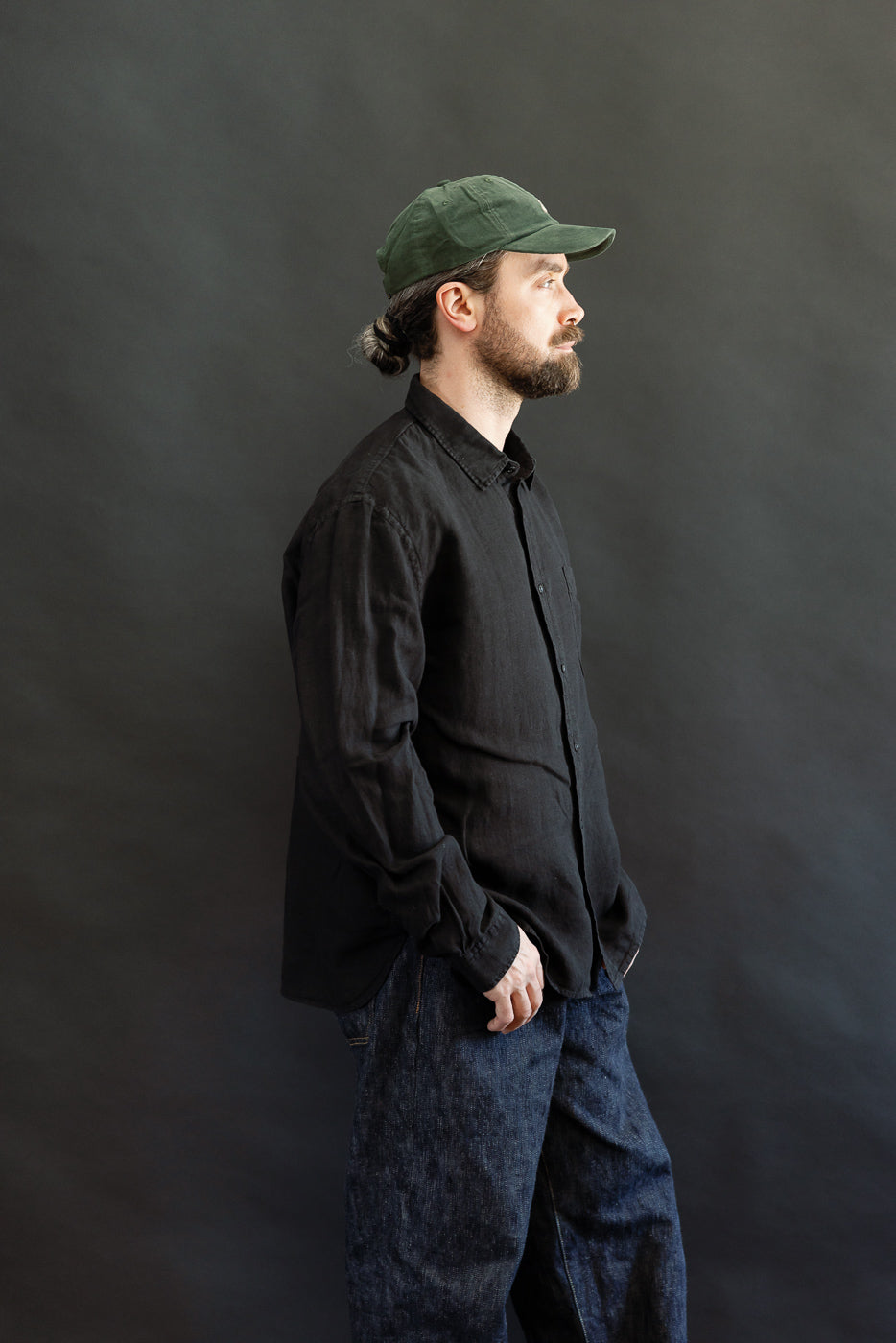 A bearded man in profile wears the SHIRT20LI.99 - Organic Linen Long Sleeve Shirt - Black, showcasing its airy, cropped fit with a classic collar, chest pocket, and Corozo buttons.