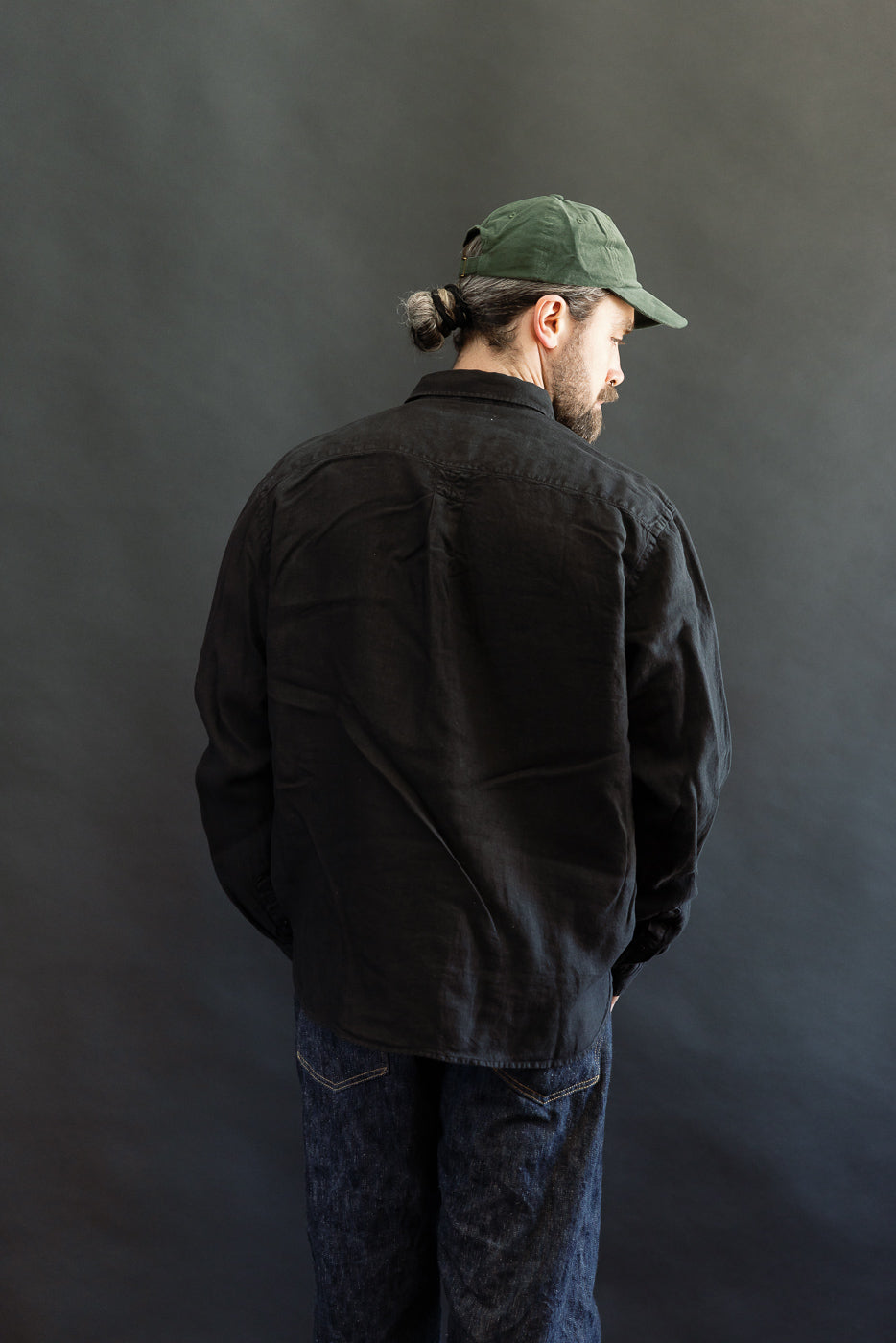 Adult male model wearing the SHIRT20LI.99 - Organic Linen Long Sleeve Shirt in black, showcasing its relaxed fit and small bun under a dark-green cap, paired with indigo denim jeans, emphasizing casual style.