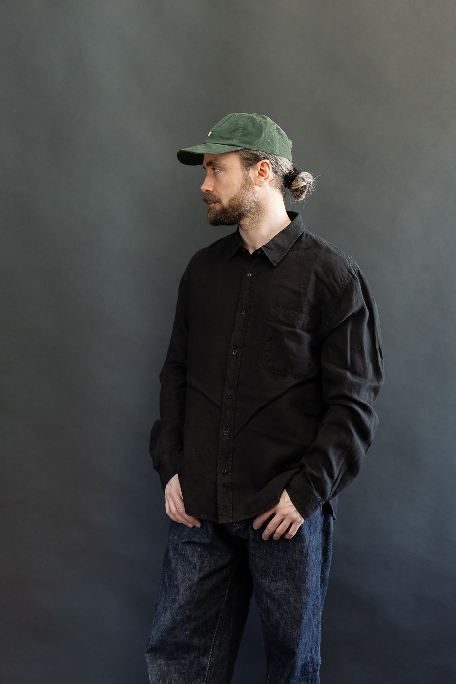 A man models the SHIRT20LI.99 - Organic Linen Long Sleeve Shirt - Black, showcasing its relaxed, cropped fit, single chest pocket, and Corozo nut buttons, embodying Dant's heritage-inspired fashion.