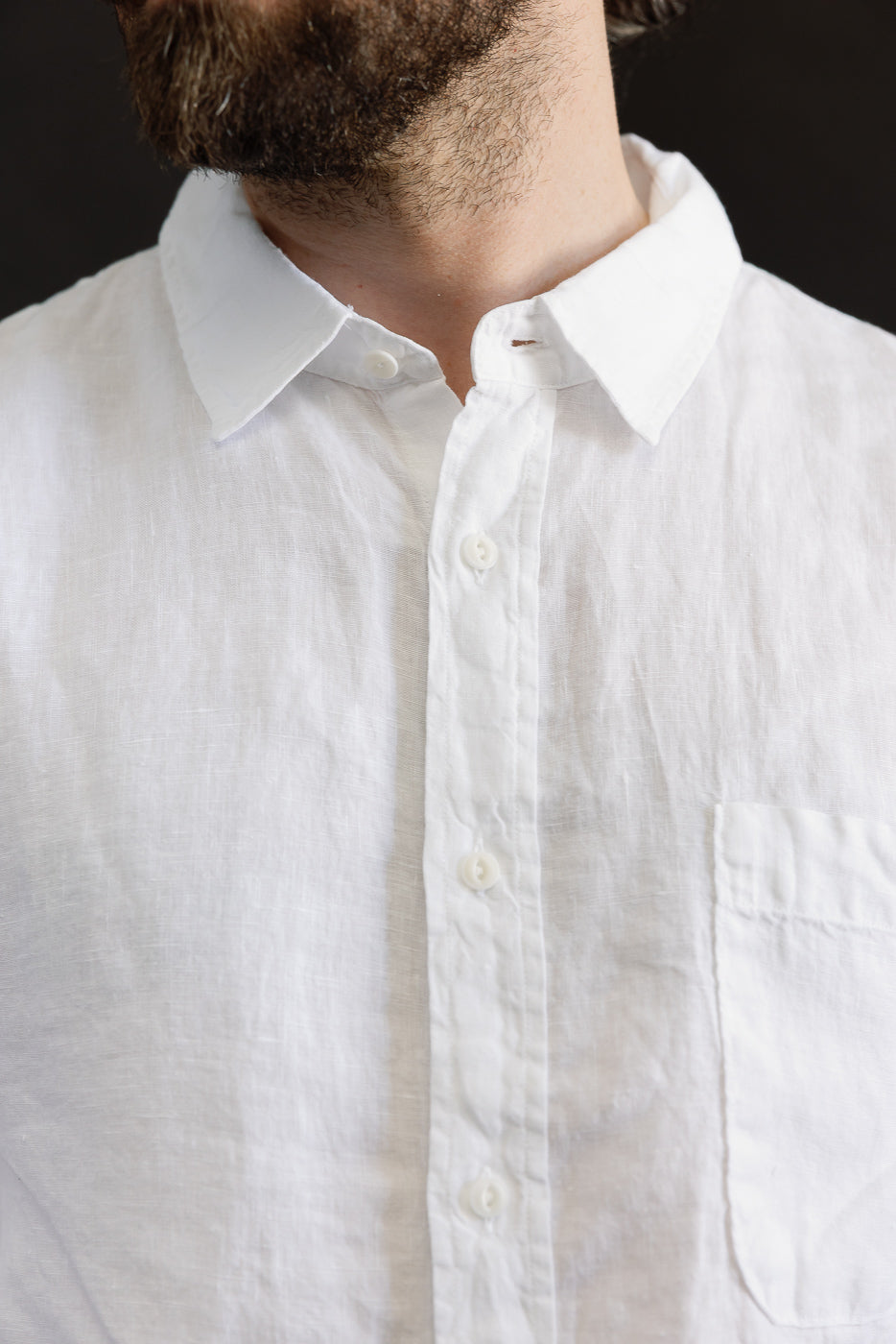 Close-up front of white organic linen long sleeve shirt on male model, showing collar, buttons, pocket, and fabric texture. Model is 5'9