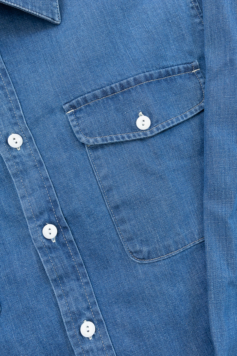 Close-up front view of a blue denim shirt showing the chest pocket, white buttons, and fabric texture.