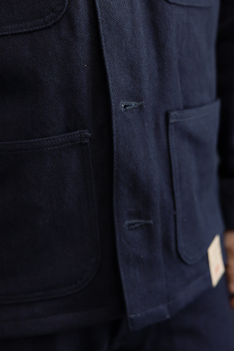 Close-up of navy work jacket front, showing textured fabric, flap pockets, buttonholes, and tag detail on male model.