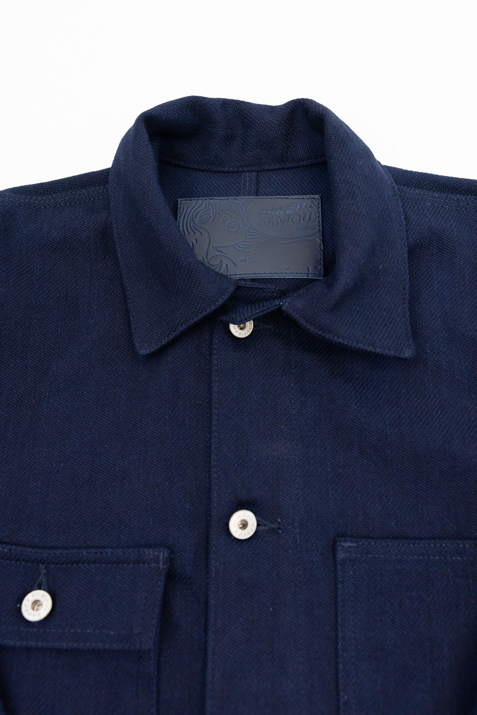 Close-up front view of navy work shirt: collar, branded tag, silver buttons, chest pockets, textured fabric