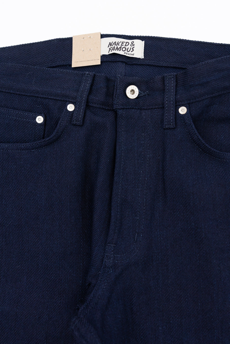 Close-up front view of dark denim jeans with Naked & Famous tag, button fly, and silver rivets