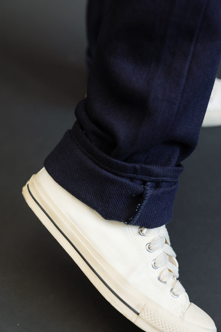 Close-up of navy selvedge denim pant cuff, cuffed hem detail, worn by male model with cream sneakers