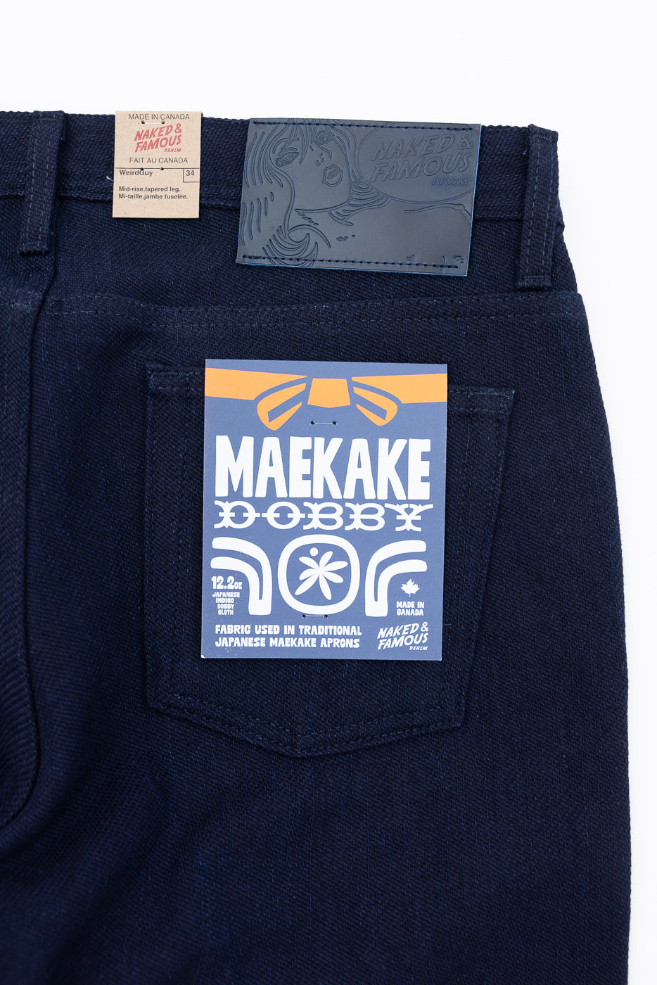 Close-up back view of dark denim jeans showing waistband tags, back pocket with Maekake Dobby label, and fabric texture