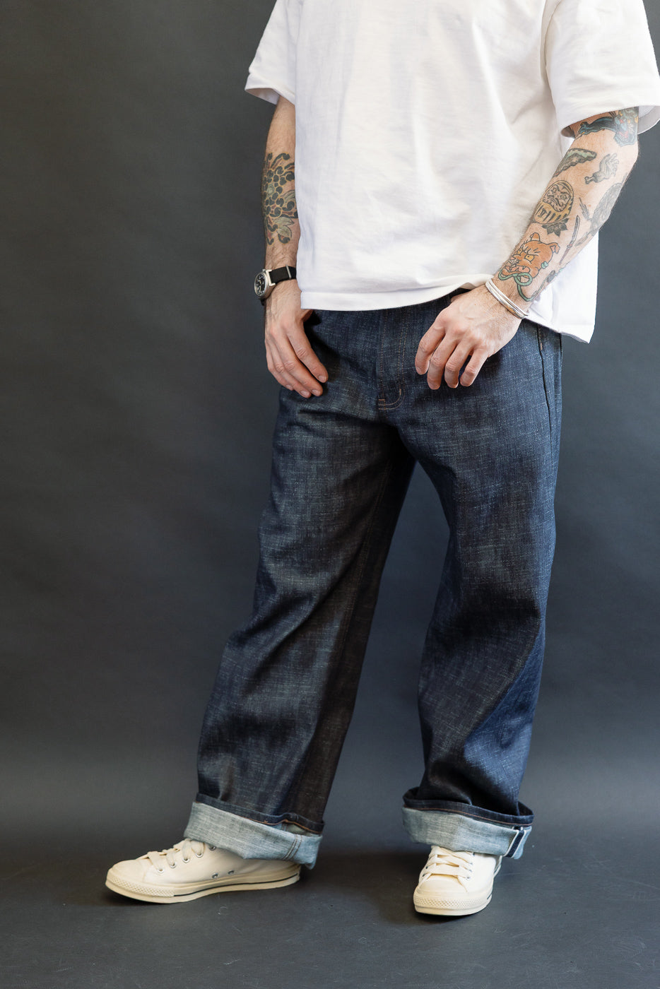 A casually dressed person models the 12.5oz Greencast Stretch Selvedge - Strong Guy jeans, showcasing the dark indigo wide-cuffed denim with off-white sneakers against a dark-gray backdrop.