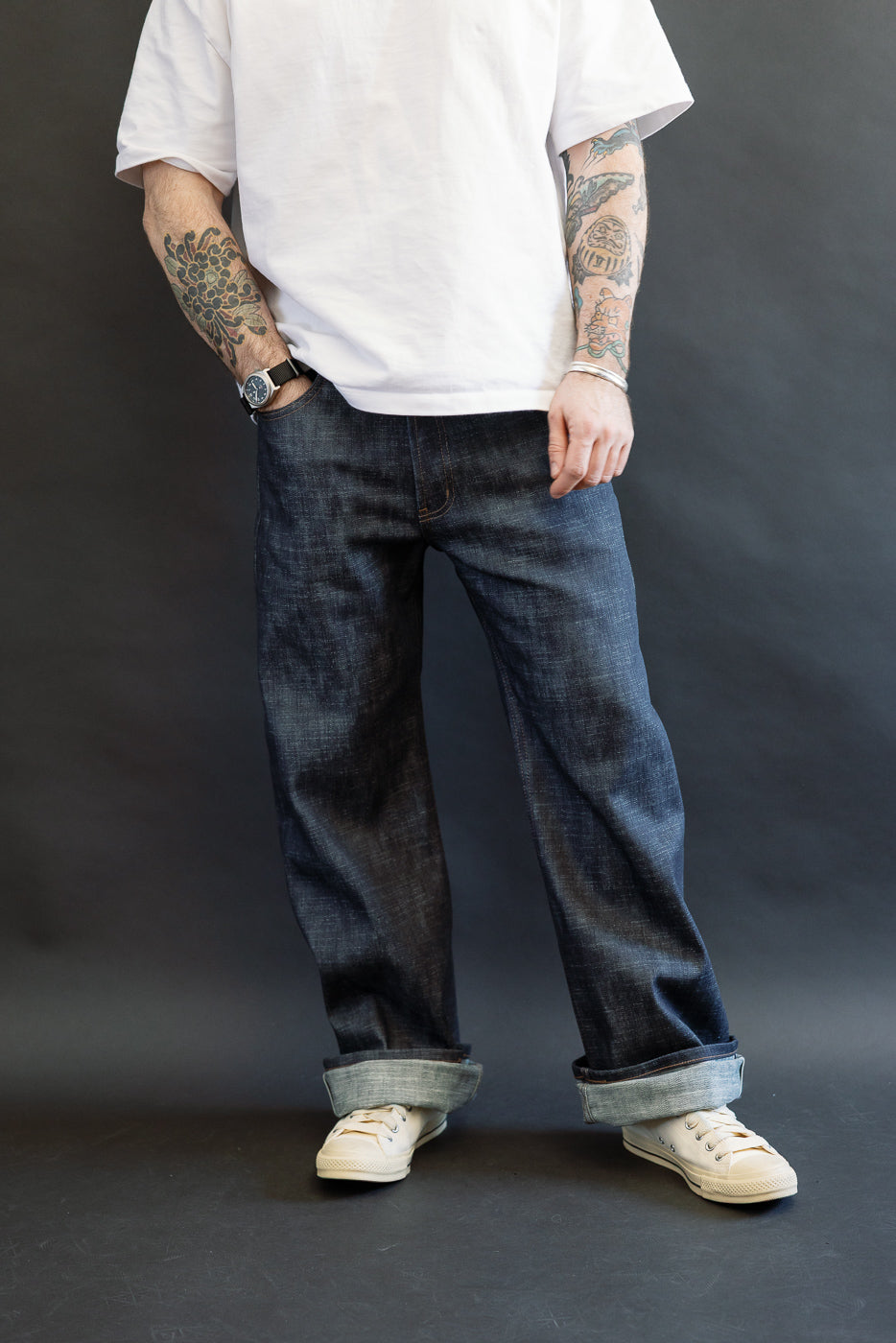 Person wearing 12.5oz Greencast Stretch Selvedge - Strong Guy jeans with wide cuffs, paired with a white T-shirt and canvas sneakers. The denim features a textured surface and subtle green cast.