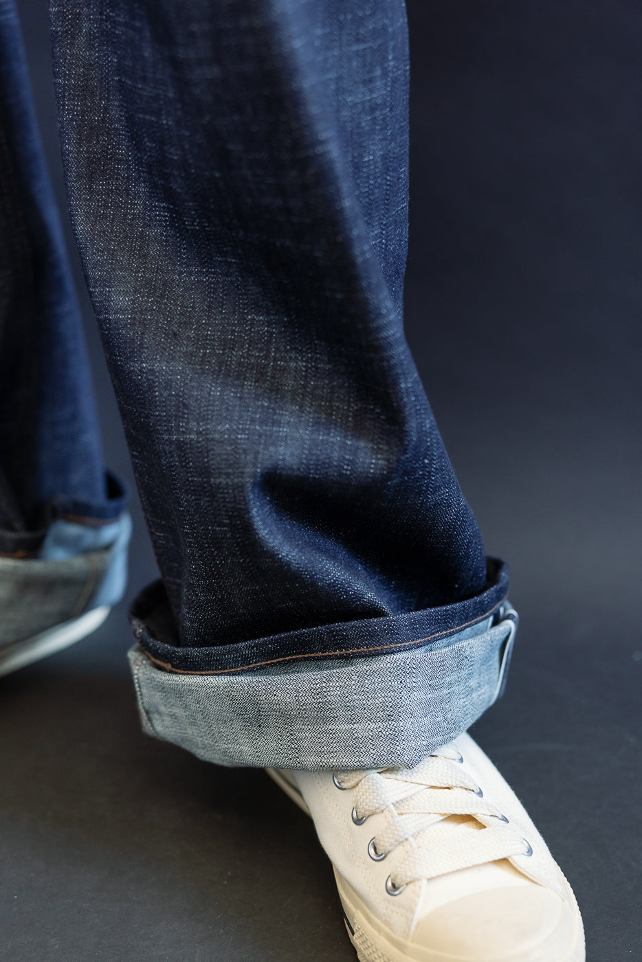 The 12.5oz - Greencast Stretch Selvedge - Strong Guy jeans, cuffed to reveal a lighter underside, paired with white canvas sneakers on a dark surface.