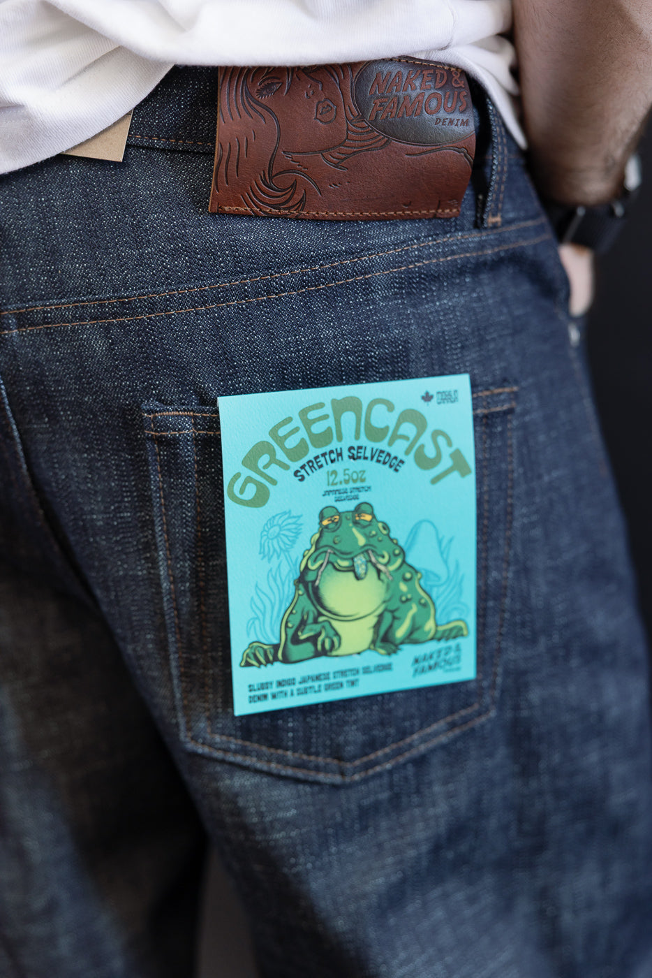 Close-up of the Naked & Famous 12.5oz Greencast Stretch Selvedge - Strong Guy jeans' back pocket, revealing a Greencast hang-tag and leather patch, worn with a white shirt.